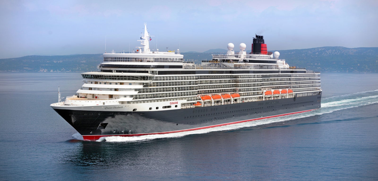 Fincantieri | Group - Business - Products - Cruise Ships - Queen Elizabeth: Side view of Queen Elizabeth cruise ship in operation navigating near coastline, multiple passenger decks, red and black details, visible lifeboats along sides, distant hills in background.