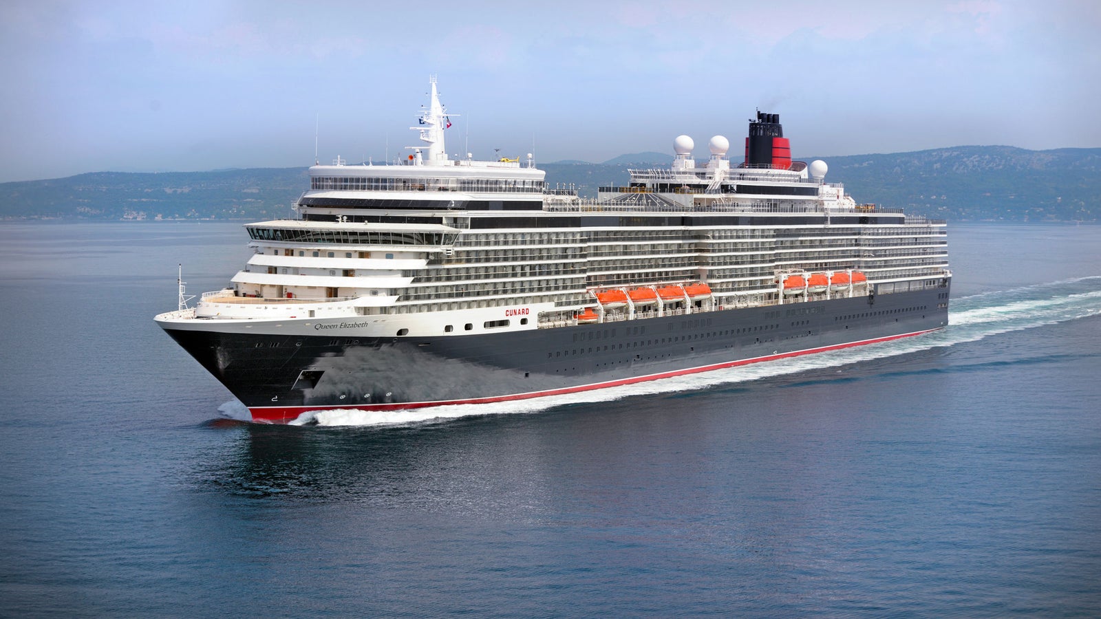 Fincantieri | Group - Business - Products - Cruise Ships - Queen Elizabeth: Side view of Queen Elizabeth cruise ship in operation navigating open sea, light hull and superstructure with red and black details, multiple passenger decks, visible lifeboats along sides, distant hills in background.