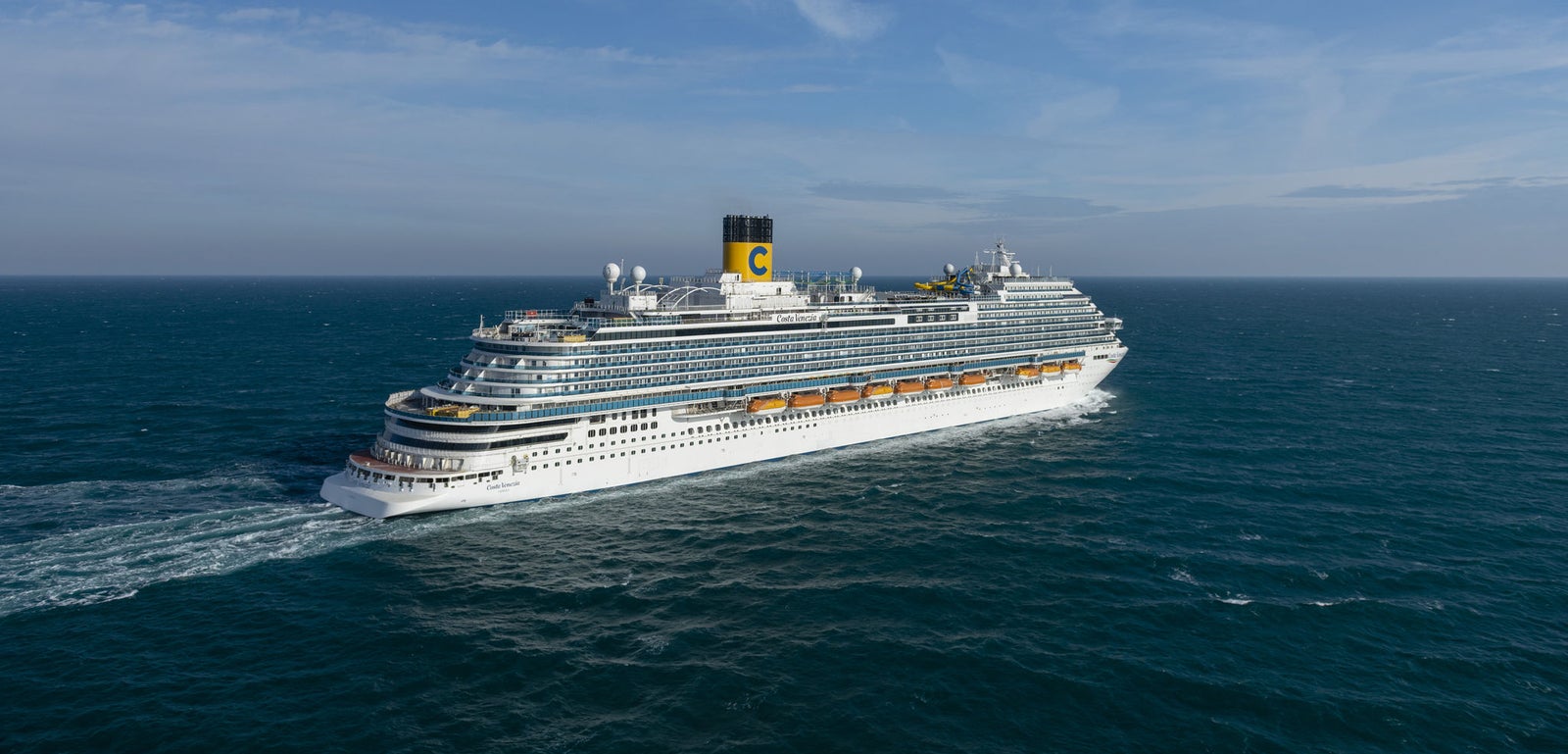 Fincantieri | Group - Business - Products - Cruise Ships - Costa Venezia: Large operational cruise ship with multiple decks, yellow funnel with 'C' logo clearly visible, sailing on calm waters under clear sky, grandeur and refined design highlighted.