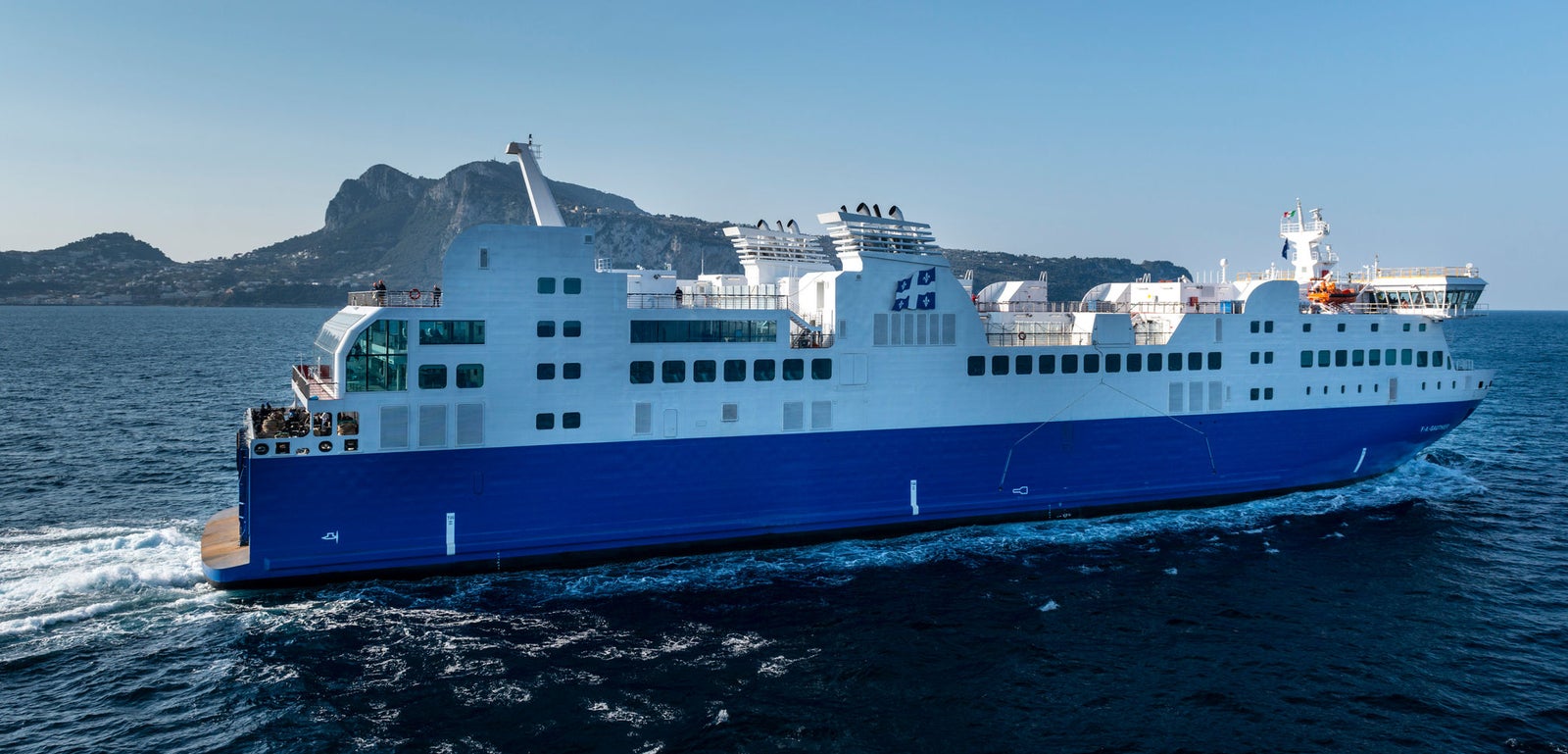 Fincantieri | Group - Business - Products - Ferries - Cruise FA Gauthier: Modern white and blue "FA Gauthier" ferry in operation, sleek design, multiple decks, visible windows, cruising through calm blue waters, scenic coast and clear sunny sky in background.