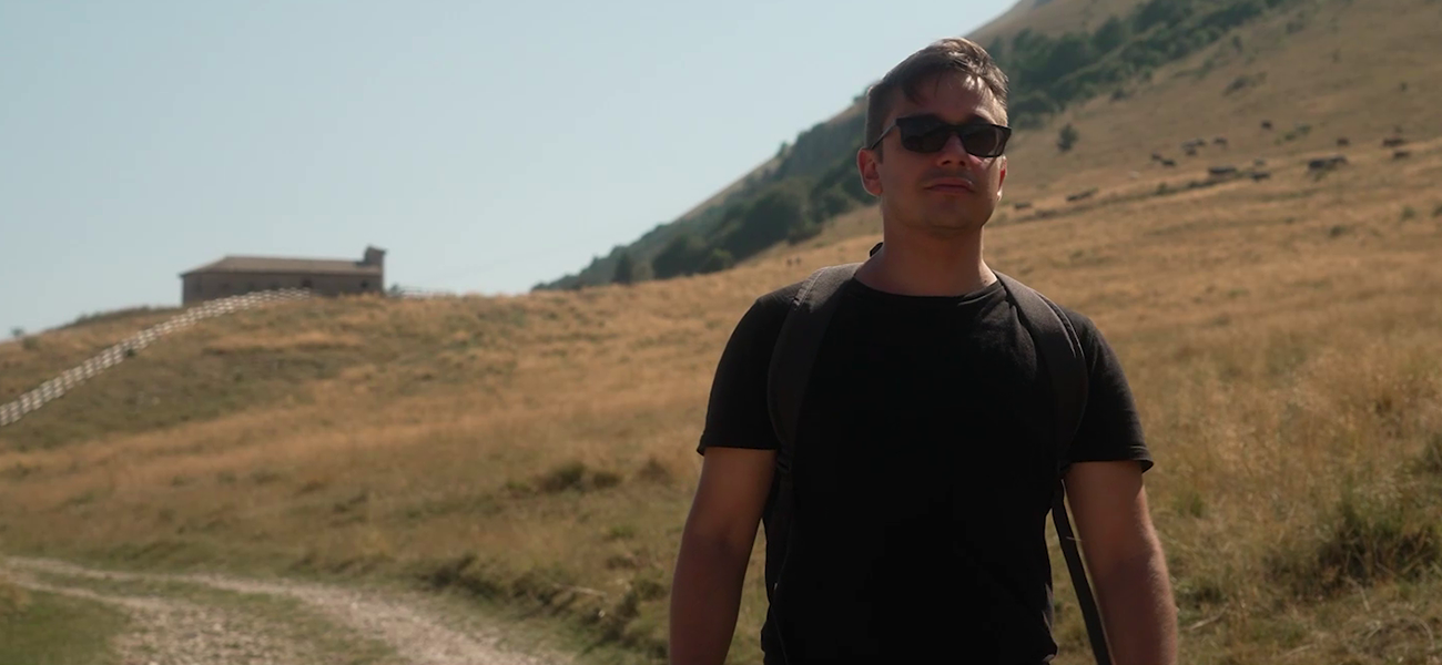 Fincantieri – People: Portrait of a young man in a black t-shirt and sunglasses, shoulder strap visible on the left shoulder, hilly background with dry vegetation and blue sky, strong sunlight, warm and natural color palette, half-length framing with blurred background to emphasize the subject.