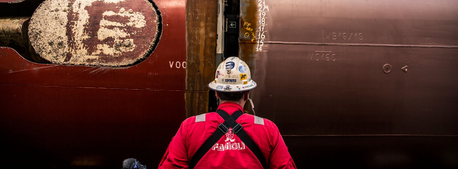 Fincantieri | Services - Ship Repair and Conversion: Operator in red uniform and helmet standing in front of a ship hull under repair, weld marks and measurement numbers (15 and 16) visible on the metal surface, industrial environment.