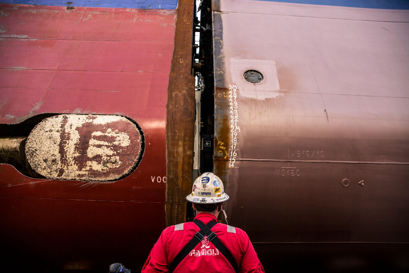 Fincantieri | Services - Ship Repair and Conversion: Operator in red uniform and helmet standing in front of a ship hull under repair, weld marks and measurement numbers (15 and 16) visible on the metal surface, industrial environment.