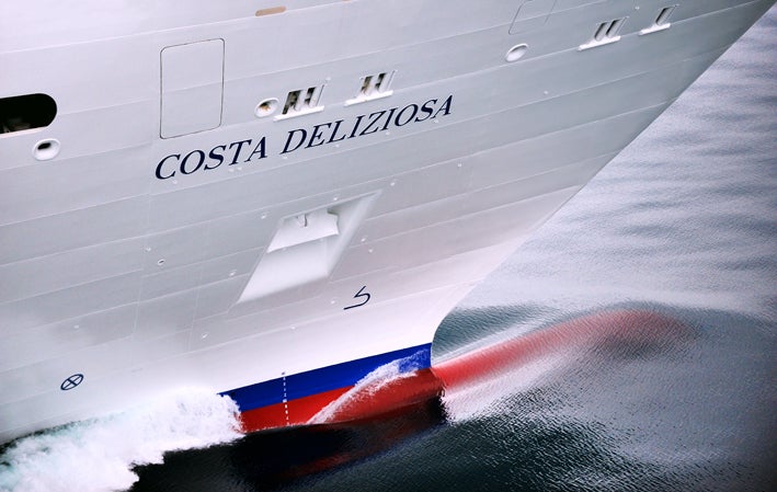 Fincantieri | Group - Business - Products - Cruise Ships - Costa Deliziosa: Bow of Costa Deliziosa cruise ship in operation cutting through calm waters, sleek design with vibrant red hull detail, ship's name prominently displayed on side, gentle waves and reflections enhance dynamic visual appeal.