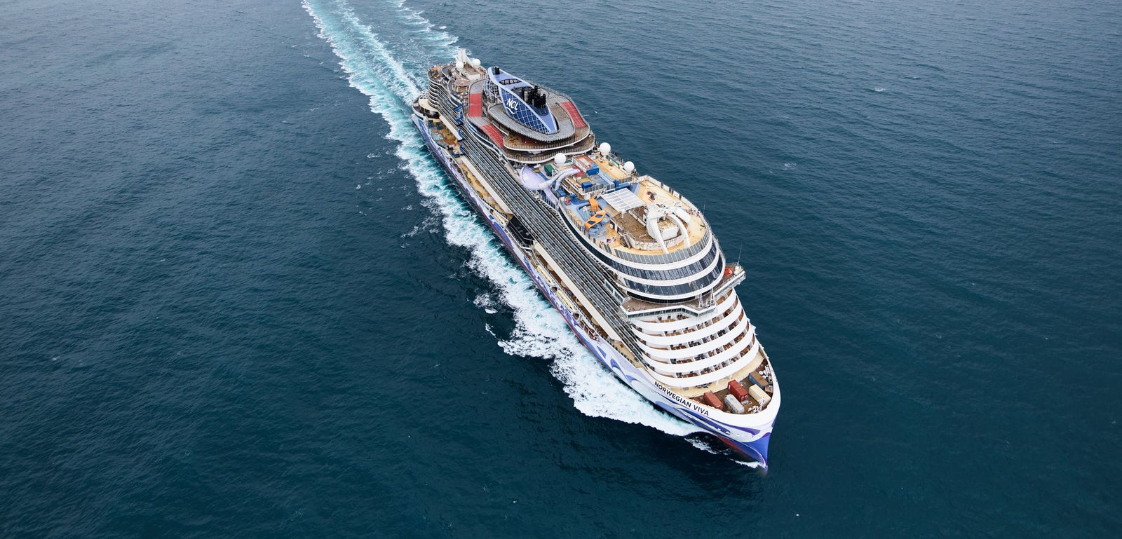 Fincantieri | Gruppo - Business - Prodotti - Navi da Crociera - Norwegian Viva: Nave da crociera Norwegian Viva operativa in navigazione su mare calmo e aperto sotto cielo nuvoloso, scafo con motivi blu e bianchi vivaci, più ponti con sedute esterne e aree ricreative, nome 'Norwegian Viva' esposto a prua.