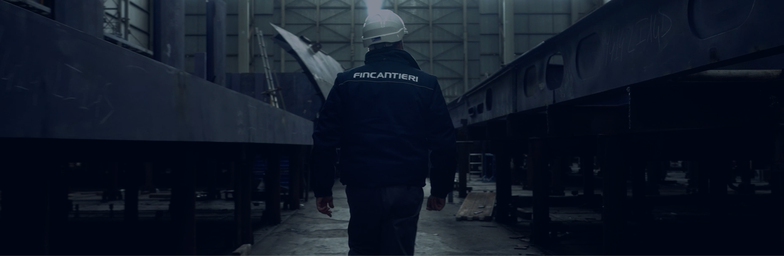 Fincantieri | People – Work With Us: Man wearing a white helmet and a jacket with "Fincantieri" written on it walks in a large industrial facility, with metal structures and soft lighting; production or shipyard context. Candidates