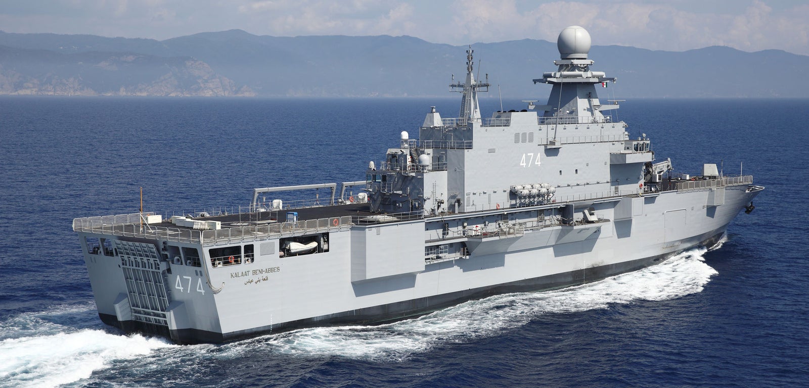Prodotto - Nave Militare Kalat Beni-Abbes: Vista laterale della nave operativa in navigazione, numero 474 visibile, ampio ponte e apparecchiature radar avanzate, sfondo montuoso.