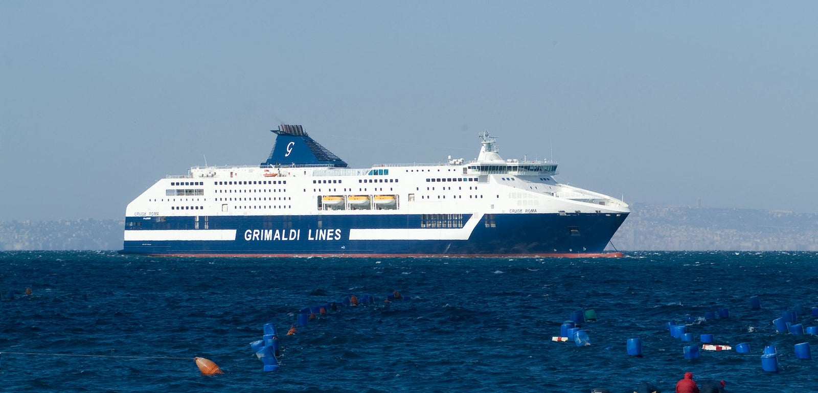 Fincantieri | Group - Business - Products - Ferries - Cruise Barcellona: Large Grimaldi Lines ferry sailing across calm blue sea, surrounded by floating buoys; small boat with two individuals in the foreground contrasts with the ferry's scale; maritime setting with clear skies and distant land on the horizon.