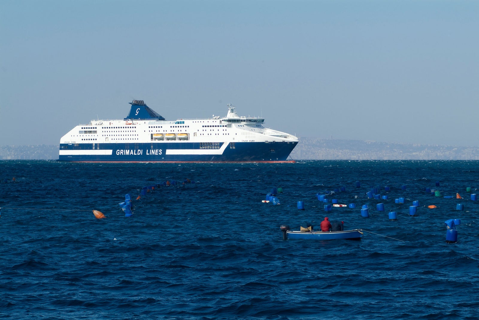 Prodotto - Traghetto Barcellona Grimaldi Lines: Vista laterale della nave operativa in navigazione su mare calmo, scafo con scritta Grimaldi Lines, presenza di boe galleggianti e piccola imbarcazione con due persone in primo piano, terra all’orizzonte e cielo sereno.