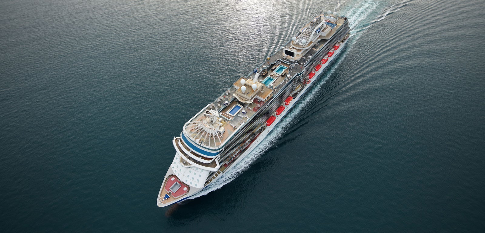 Fincantieri | Group - Business - Products - Cruise Ships - Discovery Princess: Overhead view of Discovery Princess cruise ship in operation, gliding on calm ocean waters under soft sunset sky, multiple decks, pools and recreation areas visible.