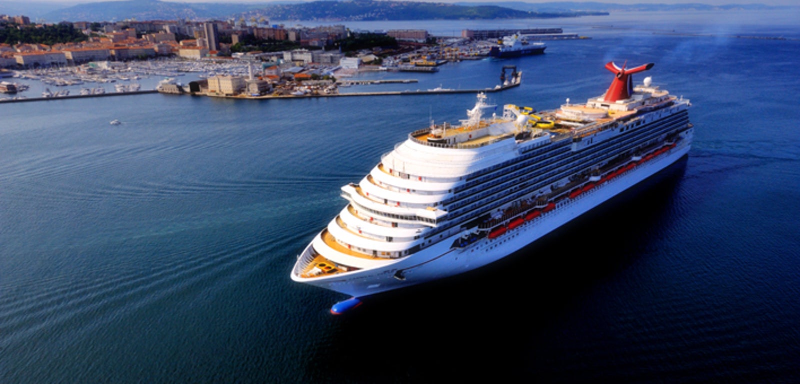 Fincantieri | Group - Business - Products - Cruise Ships - Carnival Dream: Side view of massive operational white cruise ship navigating calm blue waters near coastal city, multiple decks and distinctive red funnel, city skyline in background, serene setting emphasizes leisure travel and ocean exploration.