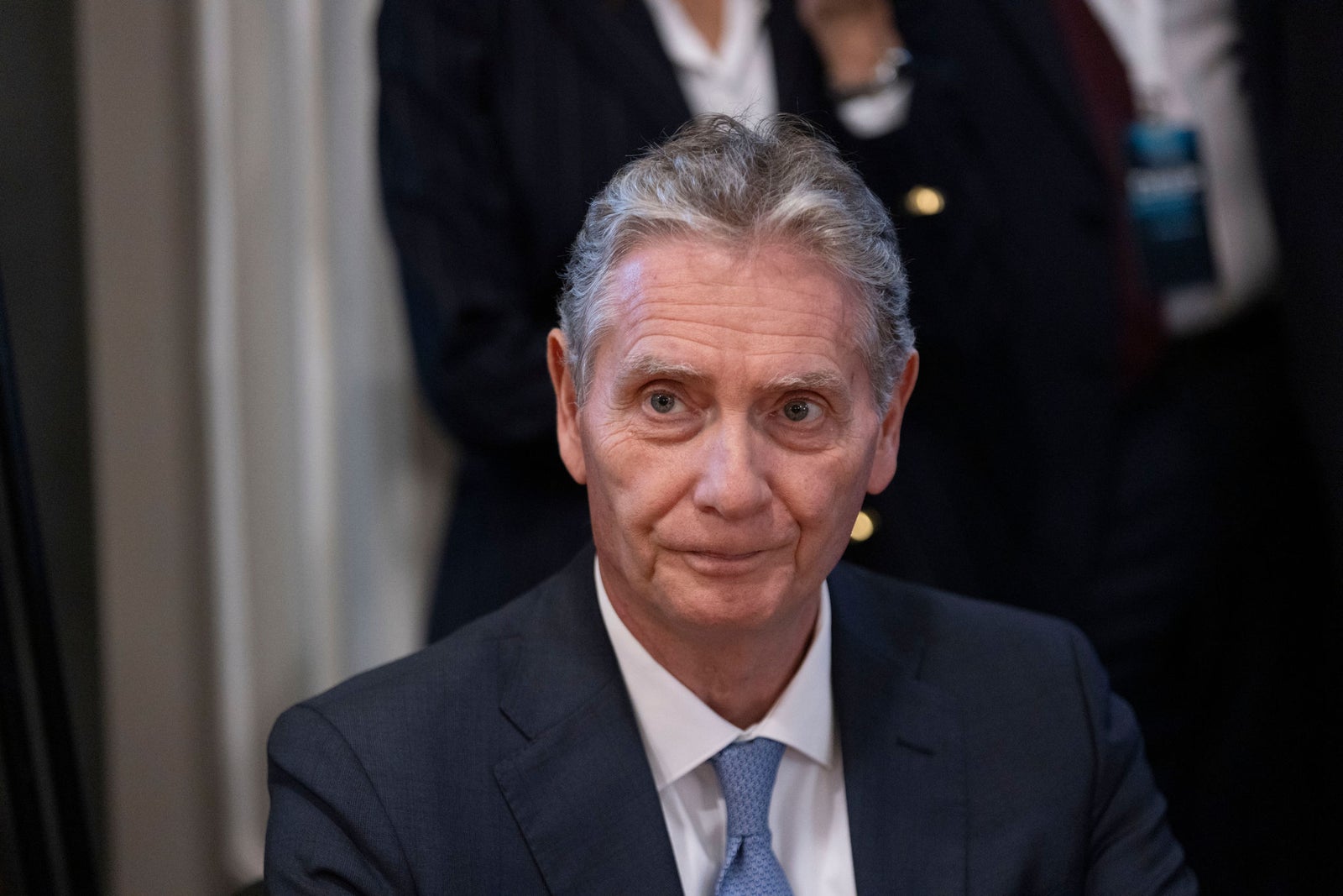Fincantieri | Newsroom, Deep: A suited individual is captured in a formal environment, wearing a dark suit and tie. The background includes blurred figures and neutral curtains, suggesting a corporate or official setting. The image conveys professionalism and a composed atmosphere.
