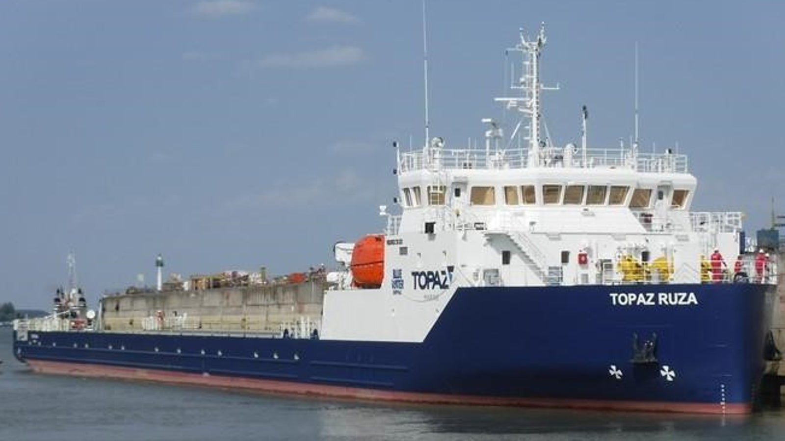 Prodotto - Nave Cargo Topaz Ruza: Vista laterale della nave ormeggiata in porto, scafo blu e bianco con testo TOPAZ RUZA visibile, design per trasporto industriale o marittimo, acque calme e cielo sereno.