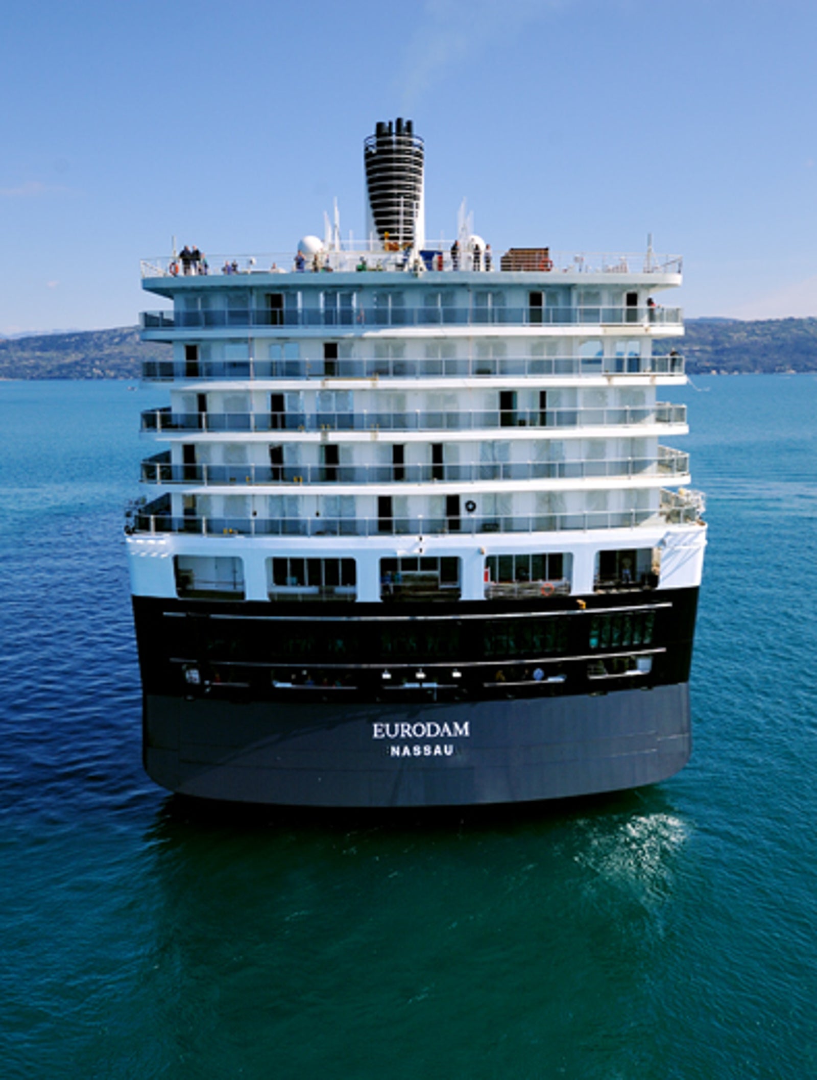  Fincantieri | Gruppo - Business - Prodotti - Navi da Crociera - Eurodam: Grande nave vista da poppa con prospettiva fisheye mentre naviga su acque calme e aperte dell’oceano, nome 'Nassau' chiaramente visibile sullo scafo, cielo azzurro luminoso e terra lontana all’orizzonte che enfatizzano l’immensità del mare.