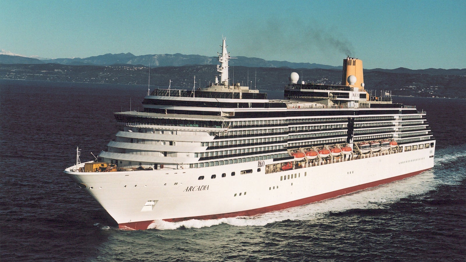 Fincantieri | Group - Business - Products - Cruise Ships - Arcadia: View of Arcadia cruise ship in operation navigating open sea with mountainous background, white hull with visible lifeboats and active funnel.