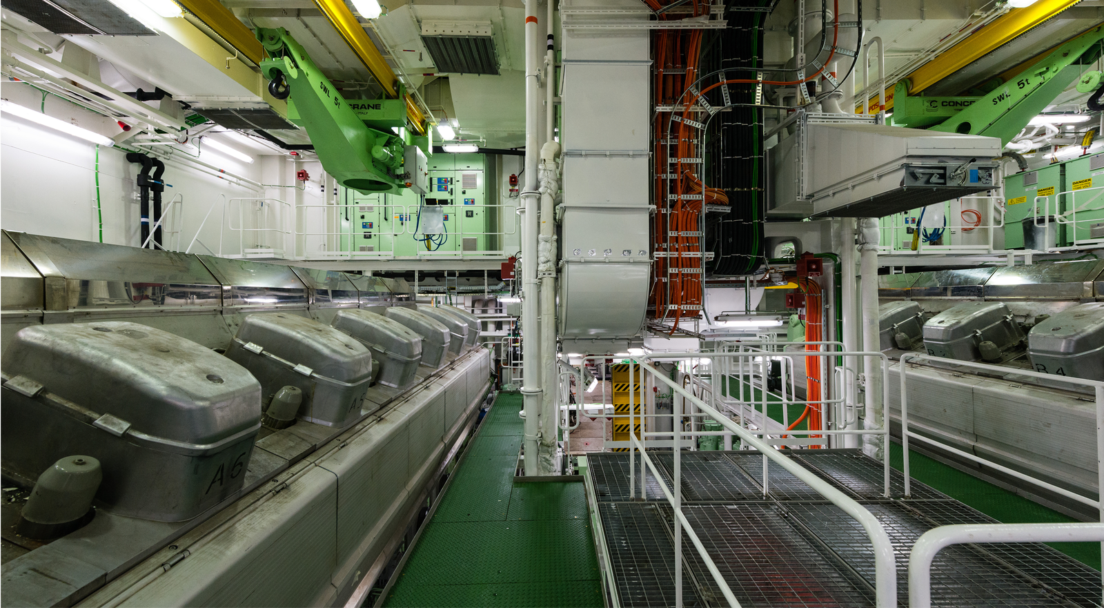Fincantieri | Group - Subsidiaries and Affiliates - Fincantieri SI: Spacious industrial engine room featuring large metallic machinery and equipment; green and white walls, overhead cranes and visible wiring; well-lit area, showcasing intricate engineering details and clean, organized layout.
