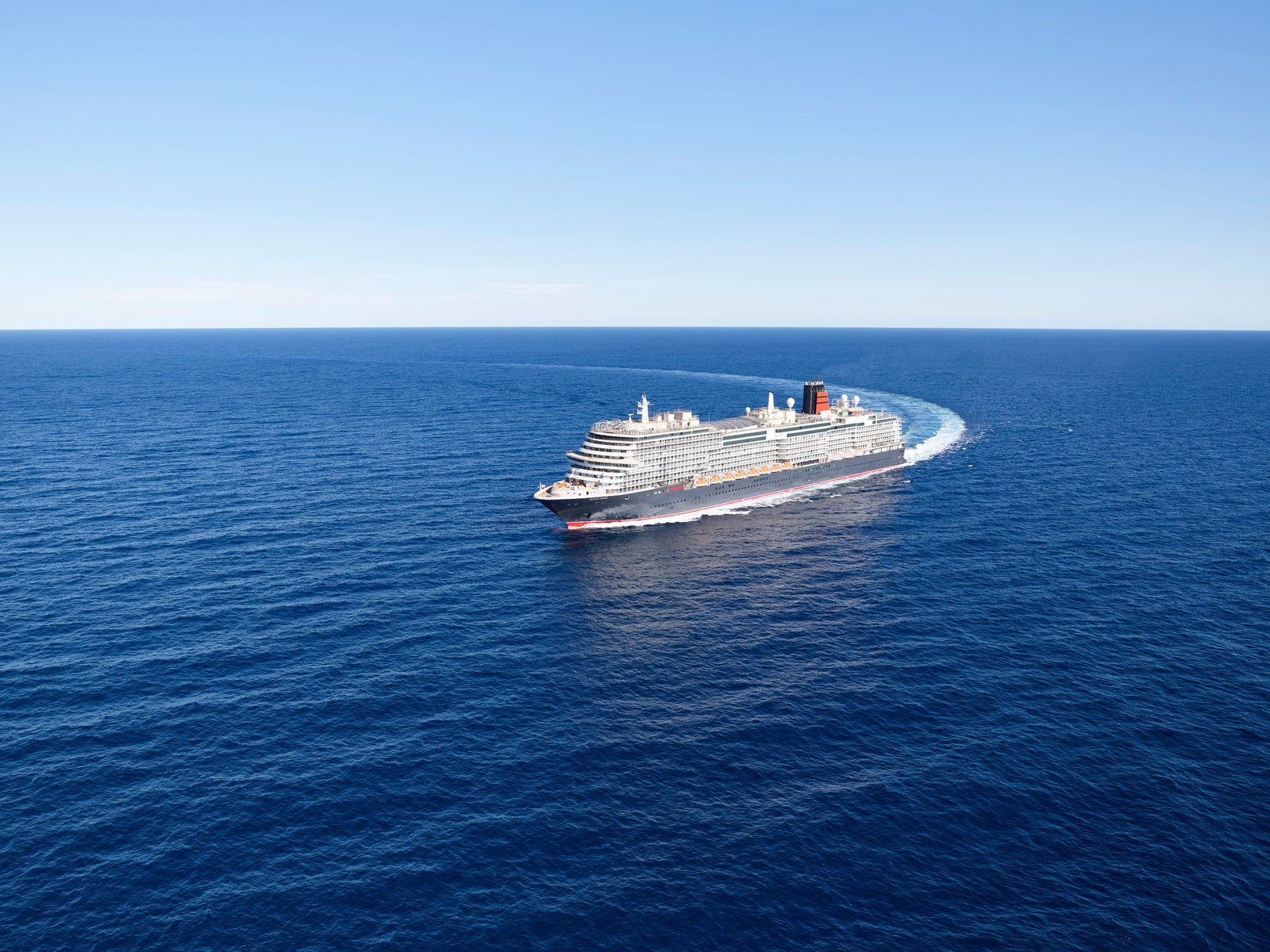 Fincantieri | Group - Business - Products - Cruise Ships - Queen Anne: Aerial side view of medium-sized cruise ship in operation, white hull with red stripe along waterline, sailing on deep calm blue sea, light wake, blue sky and sharp horizon.