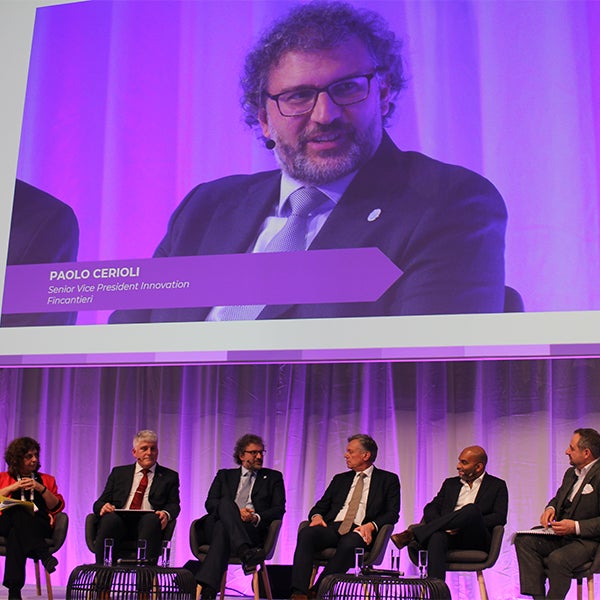 Fincantieri | Group - Innovation - Naval Ecosystem of the Future - Wave 2 the Future: Group of professionals seated on stage during a corporate panel, large screen displaying ‘PAOLO CERIOLI, Senior Vice President Innovation, Fincantieri’, purple lighting, formal and engaging atmosphere.