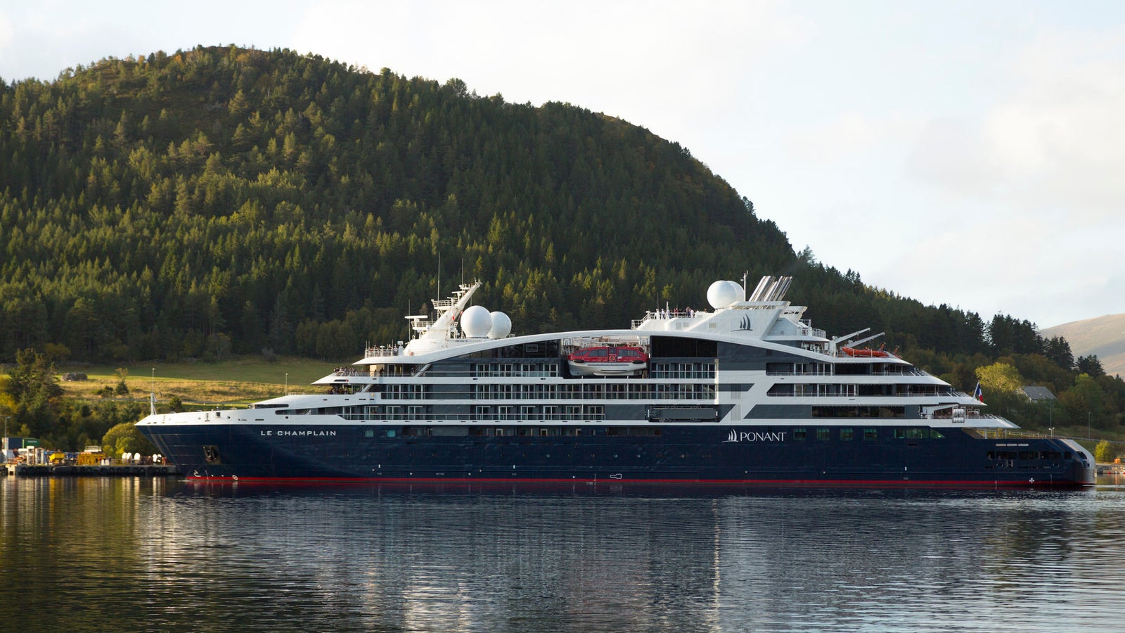 Fincantieri | Gruppo - Business - Prodotti - Navi da Crociera - Le Champlain: Vista laterale della nave Le Champlain ormeggiata vicino alla costa, scafo dal profilo lineare, scritte "Le Champlain" visibili sullo scafo, cupole satellitari visibili, sfondo colline boscose.