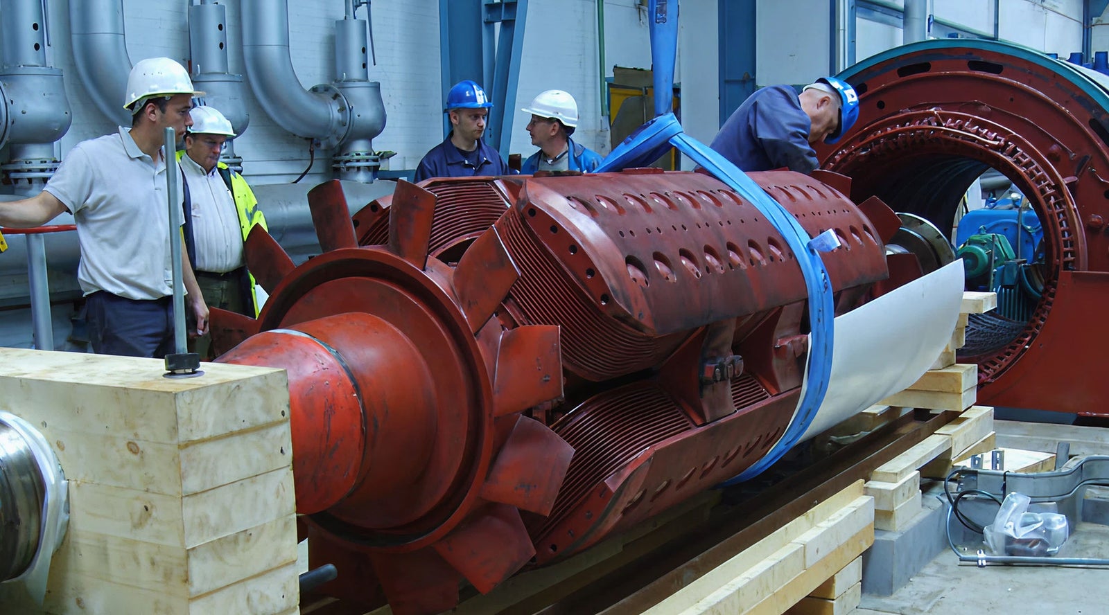 Fincantieri | Group - Subsidiaries and Affiliates - Team Turbo Machines: Large industrial turbine being assembled in factory environment, workers wearing safety helmets and reflective vests inspecting the equipment; red metallic components, secured with blue straps on wooden base, background includes industrial pipes and machinery, emphasizing manufacturing context.