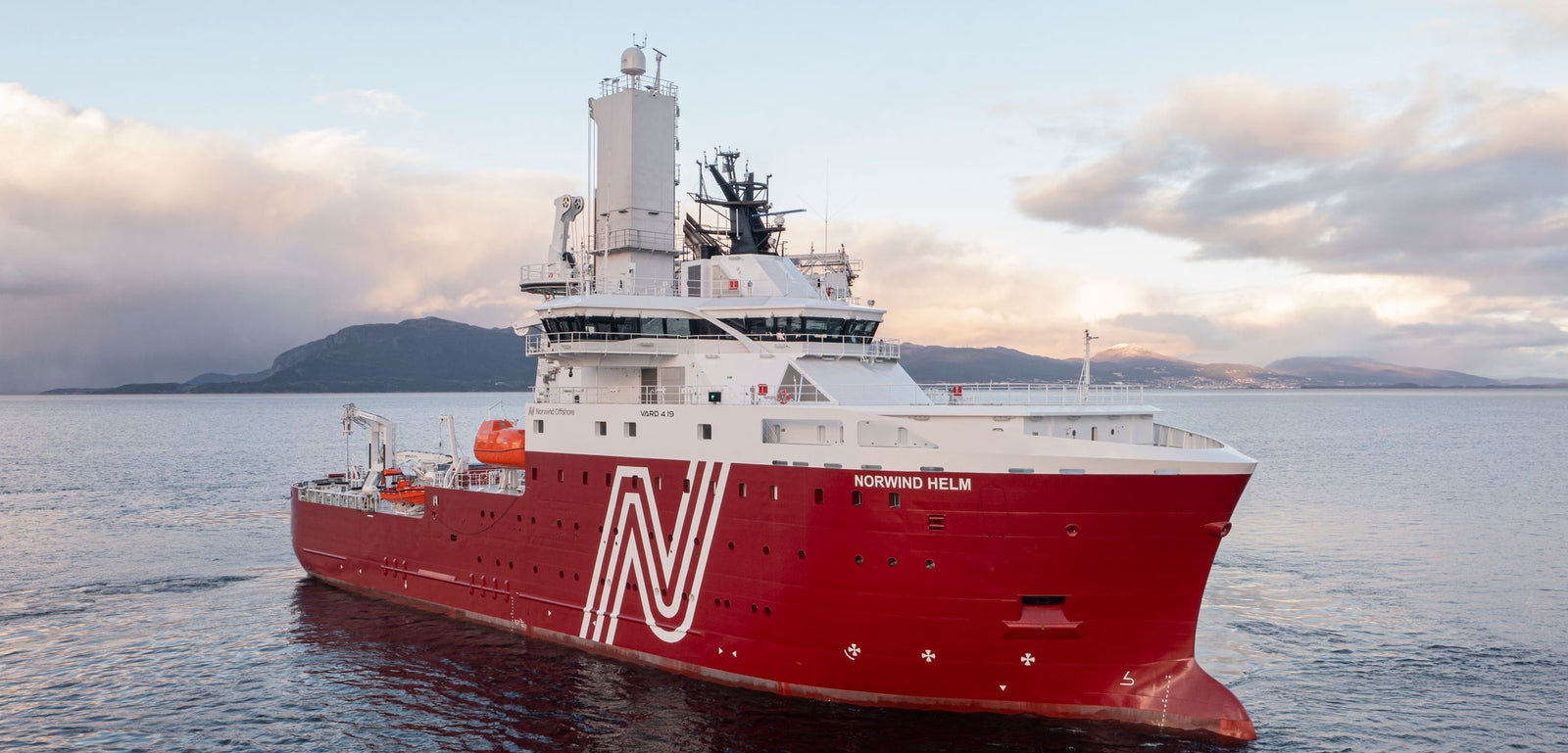 Prodotto - Offshore Support Vessel Norwind Helm: Vista laterale della nave operativa in navigazione, scafo rosso con logo N bianco e nome Norwind Helm visibile, antenne e scialuppe, sfondo montuoso e cielo nuvoloso.