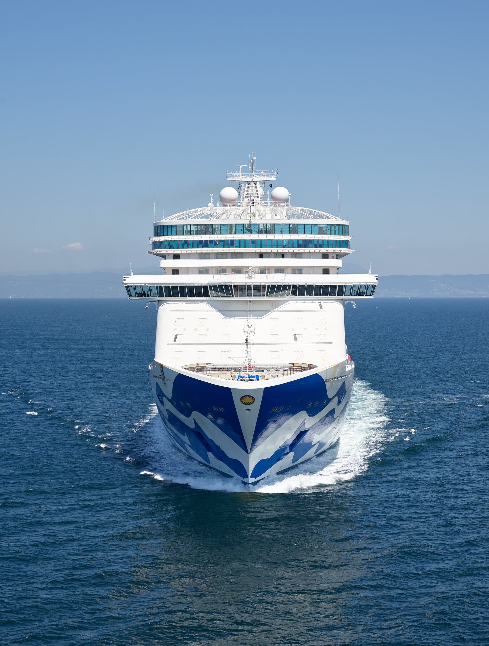 Fincantieri | Group - Business - Products - Cruise Ships - Enchanted Princess: Front view of Enchanted Princess cruise ship in operation, sailing on calm open waters under clear blue sky, wave design on hull and multiple decks visible.