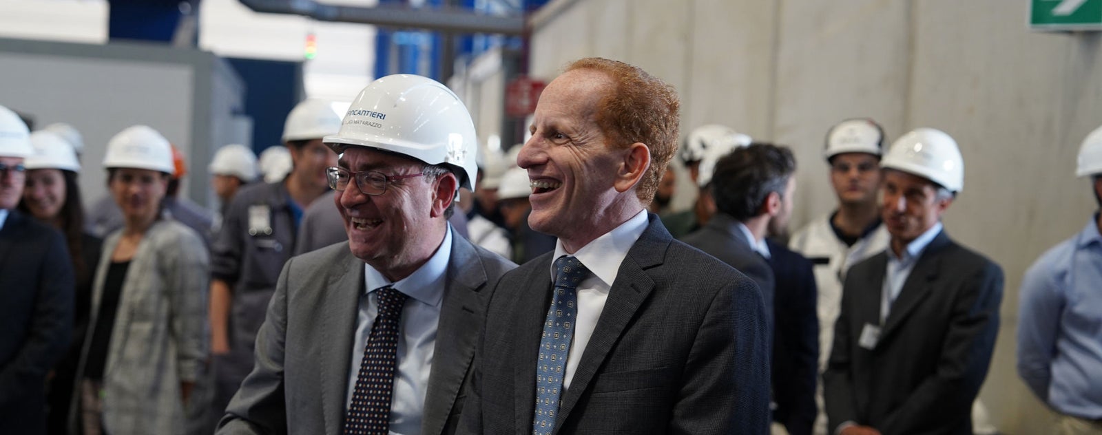 Fincantieri | Gruppo - Sostenibilità - Persone e comunità - Diritti umani: Gruppo di persone in abiti formali e caschi di sicurezza riunite in ambiente industriale, contesto di visita o ispezione professionale con sfondo di pareti in cemento e attrezzature industriali, attenzione su interazione e abbigliamento dei partecipanti.