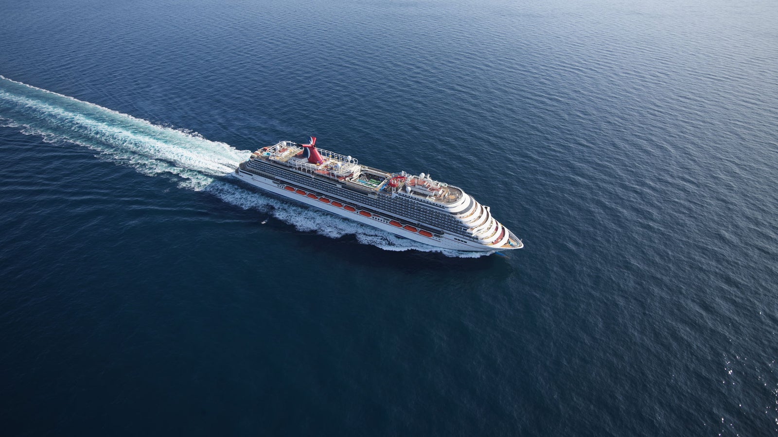  Fincantieri | Gruppo - Business - Prodotti - Navi da Crociera - Carnival Panorama: Grande nave da crociera operativa che scivola su oceano vasto e calmo sotto cielo azzurro limpido, più ponti, accenti rossi vivaci a contrasto con acqua blu intenso, scia visibile, ambiente sereno che evoca avventura e relax.