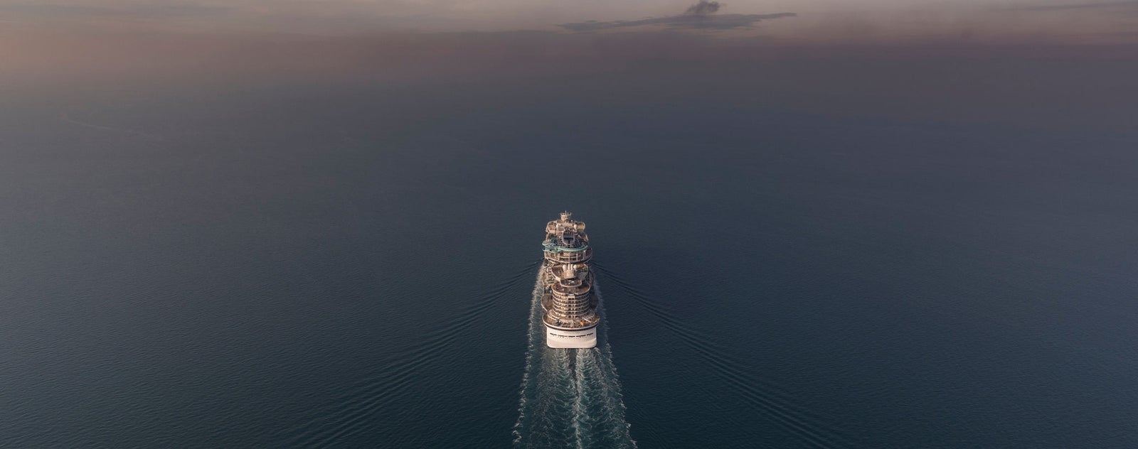 Fincantieri | Gruppo - Sostenibilità - Eccellenza industriale - Catena di fornitura: Grande nave da crociera operativa in navigazione su acque calme al tramonto, scia visibile e orizzonte sereno sullo sfondo.