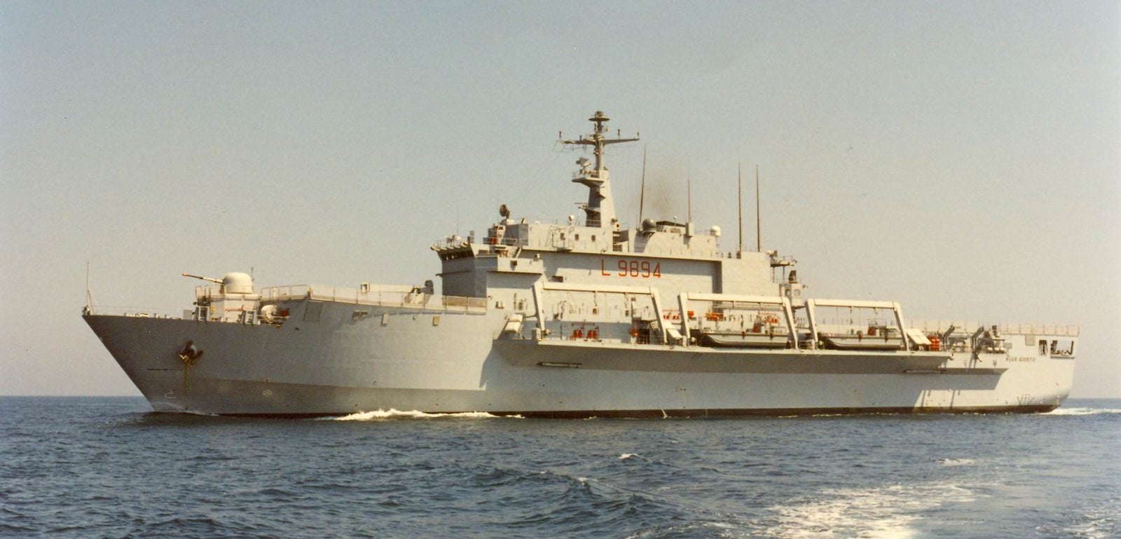 Fincantieri | Gruppo - Business - Prodotti - Navi Militari - Portaerei e Navi Anfibi - Classe San Giusto: Grande nave militare grigia in operazione, antenne e attrezzature sul ponte visibili, "999" prominente sulla struttura, in navigazione su acque calme, cielo limpido, scia visibile dietro la nave.