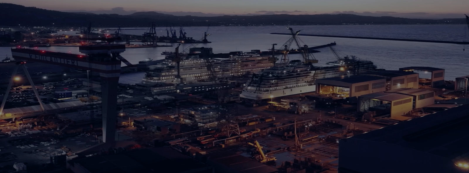 Fincantieri | Group - Business - Products - Bridges, Viaducts, Airports, Ports - Shipyard at dusk: Bustling shipyard at dusk with multiple docked vessels and cranes; artificial lights illuminate industrial activity, calm waterfront and distant hills under fading evening sky.