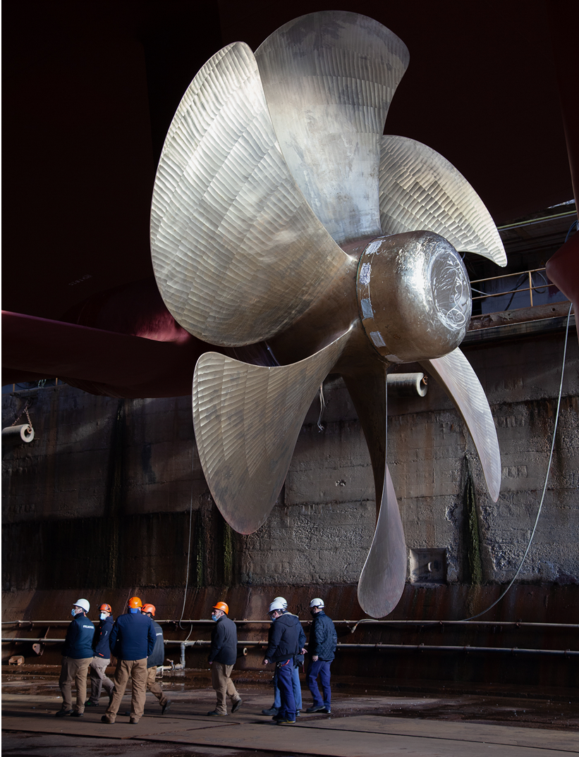 Fincantieri | Group - Sustainability - People and Community - Our People: Large metal ship propeller in a dry dock, workers with helmets around in an industrial environment with worn walls and structures, engaged in inspection or discussion, emphasis on size and technical precision.