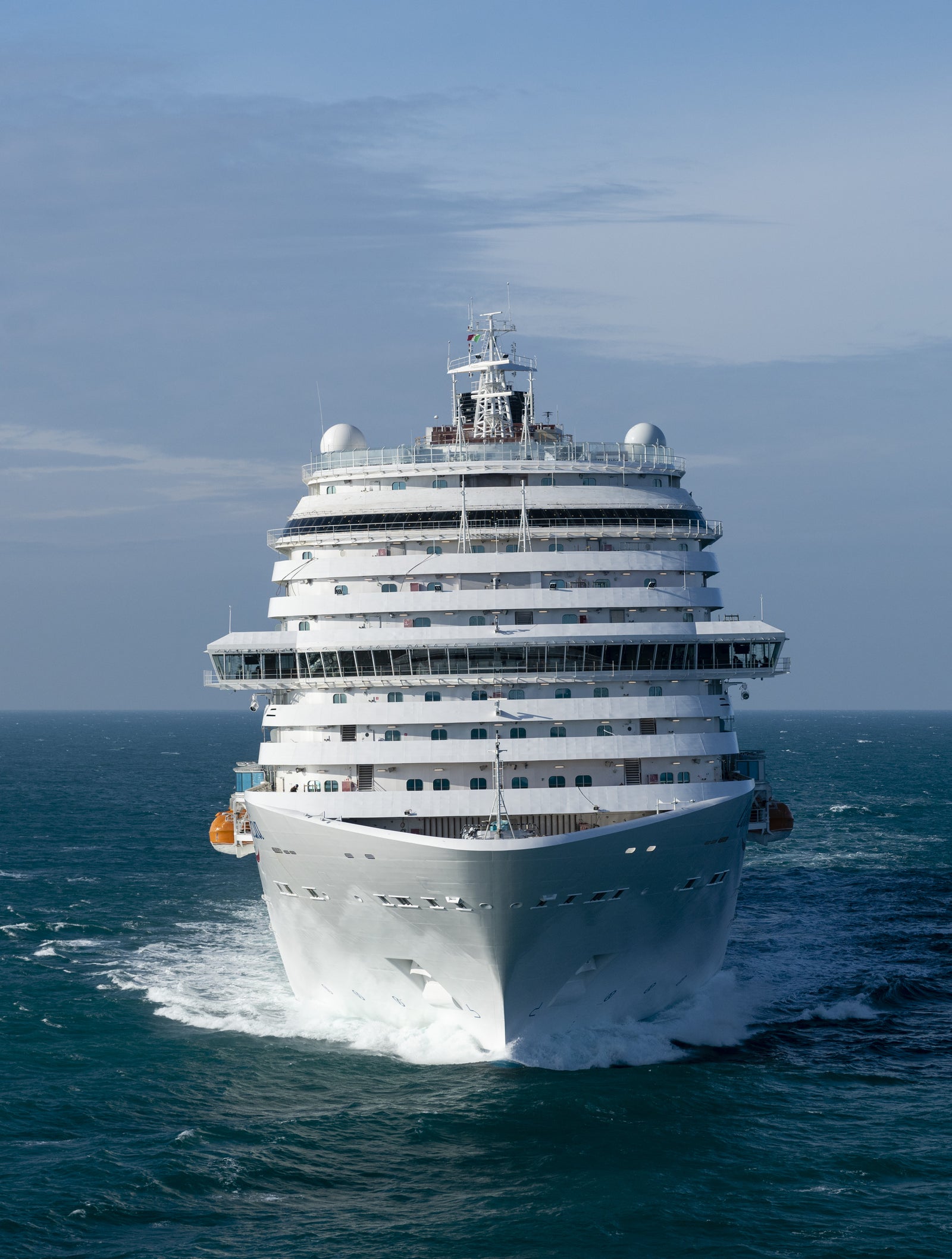 Fincantieri | Group - Business - Products - Cruise Ships - Costa Venezia: Large operational white cruise ship with many decks and distinctive yellow funnel with 'C' logo, sailing on calm waters under clear sky, modern design, grandeur and tranquil atmosphere of adventure travel.