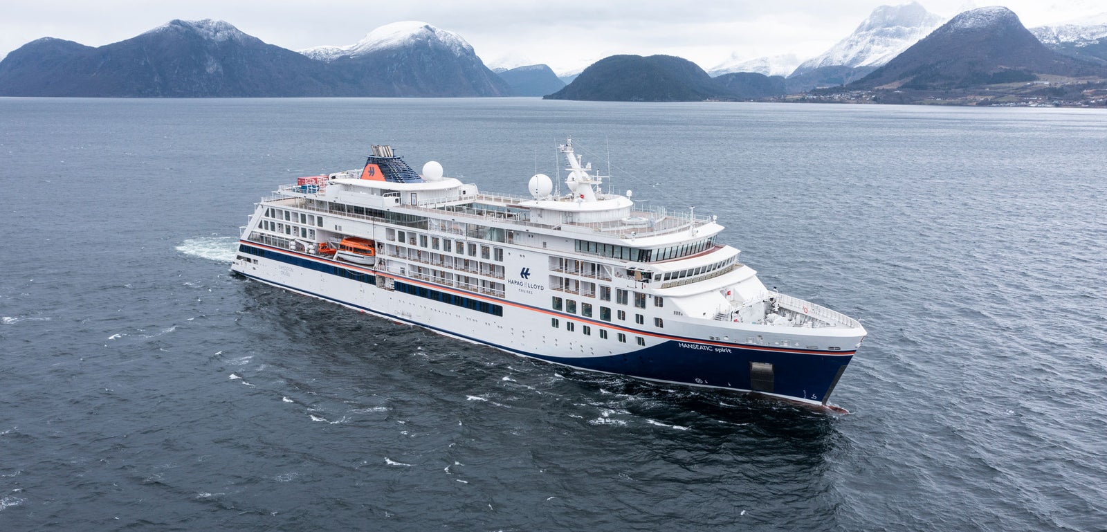 Fincantieri | Group - Business - Products - Cruise Ships - HANSEATIC spirit: Modern cruise ship in operation sailing through calm Nordic waters, surrounded by snow-capped mountains and rugged landscapes, sleek design with multiple decks and visible lifeboats, overcast sky adds serene and dramatic mood.