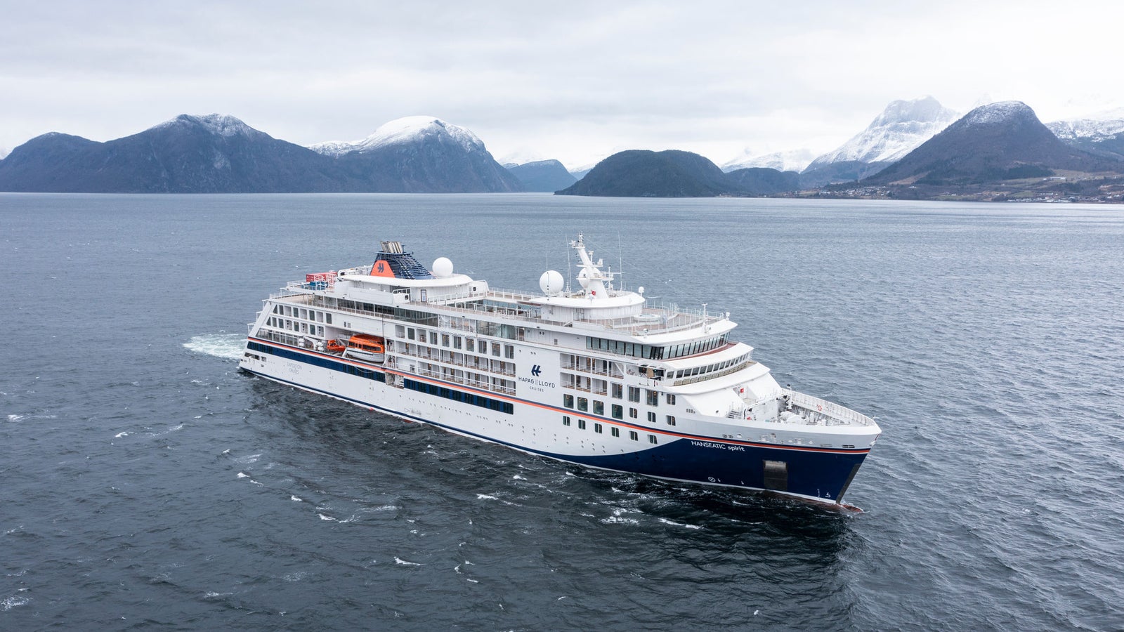 Fincantieri | Group - Business - Products - Cruise Ships - HANSEATIC spirit: Modern cruise ship in operation sailing through calm Nordic waters, surrounded by snow-capped mountains and rugged landscapes, sleek design with multiple decks and visible lifeboats, overcast sky adds serene and dramatic mood.