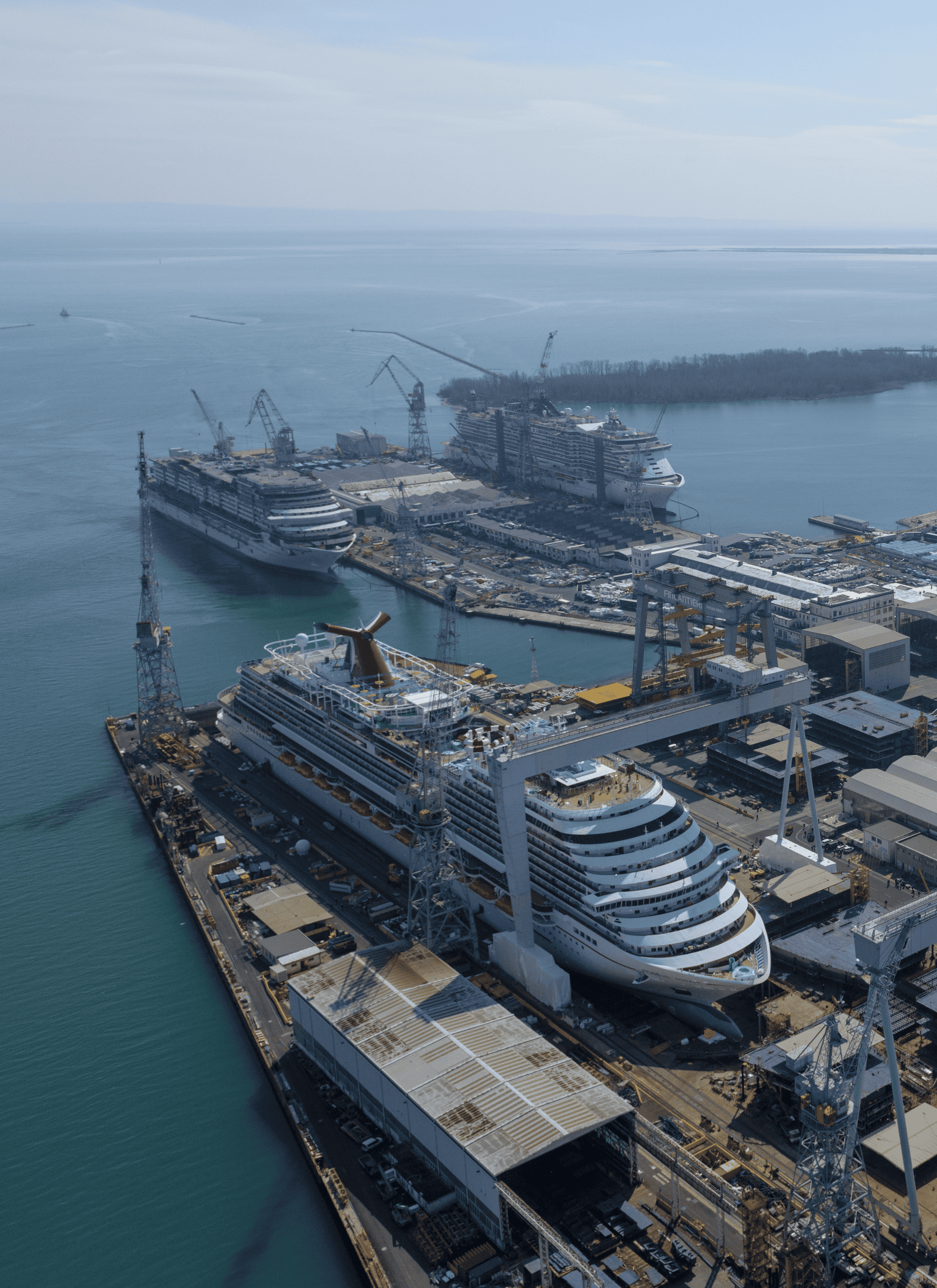 Fincantieri | About Us - Global Presence: Large shipyard with multiple cruise ships docked and industrial structures, coastal area with cranes and maritime infrastructure, clear day with a view of the water and surrounding landscape.