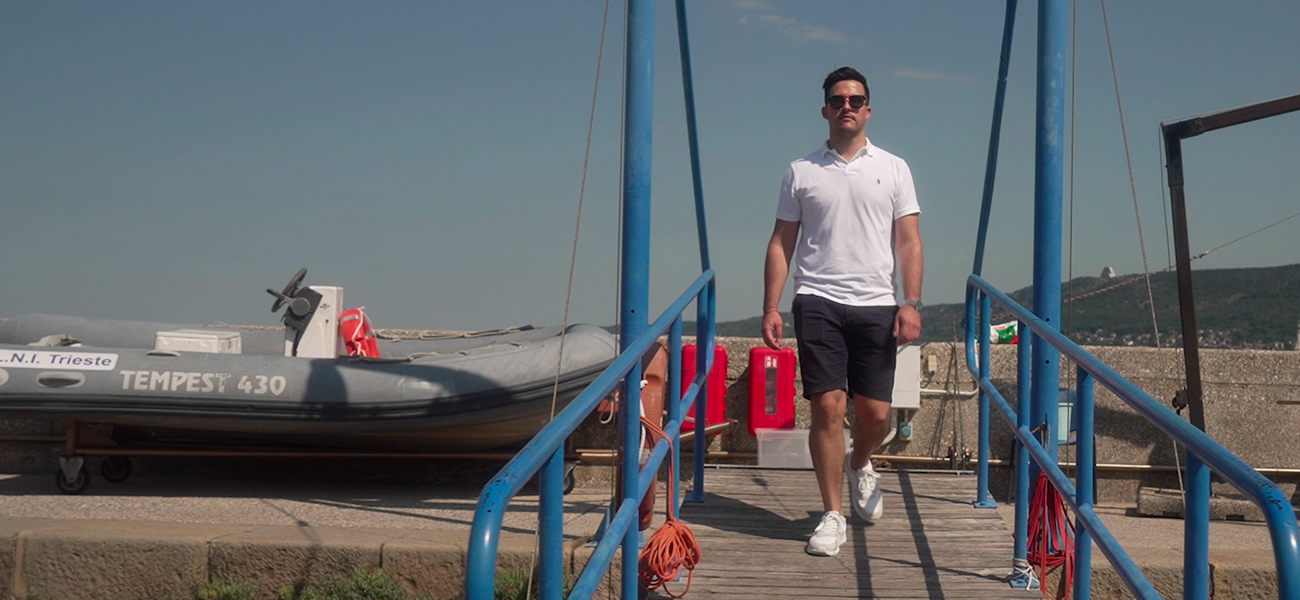 Fincantieri – People: Man in casual clothing walking on a pier with blue handrails on a sunny day; in the background, a Tempest 430 inflatable boat and the inscription “N.I. Trieste”; marina or port area setting with clear sky.