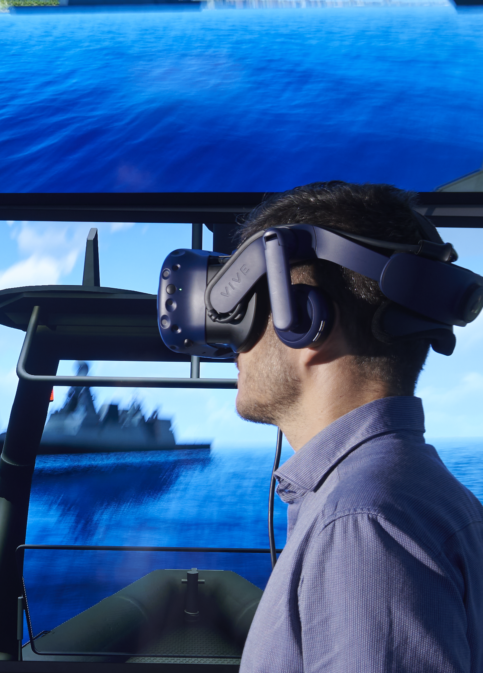 Fincantieri | People - Innovative Spirit: Man wearing a virtual reality headset uses a maritime simulator, two screens showing ocean, ships, and coasts, control panel with steering wheel and numerical indicators, immersive experience.