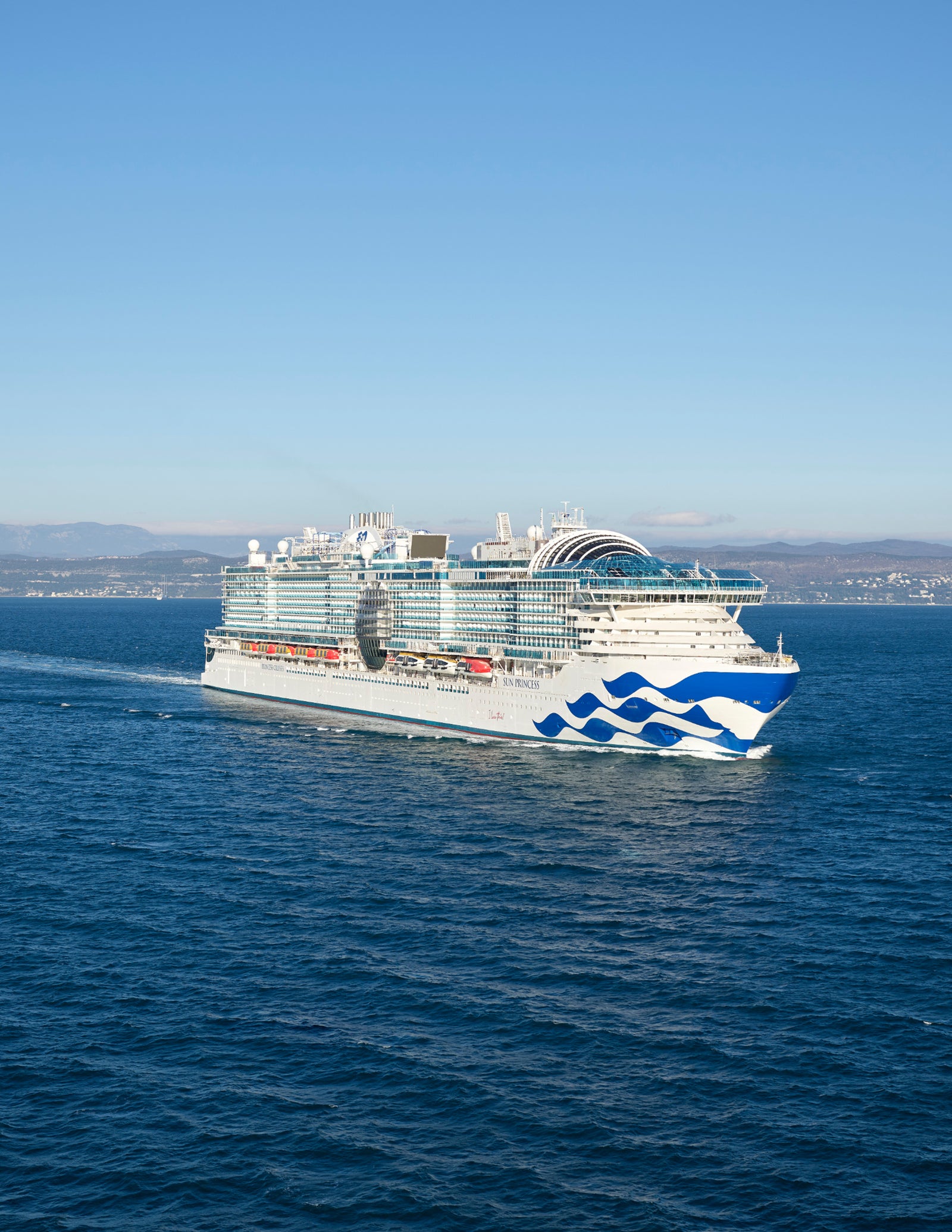 Fincantieri | Sostenibilità - Transizione Energetica: Large white cruise ship with blue details operationally sailing on the open sea under a clear sky, visible wake behind the ship.