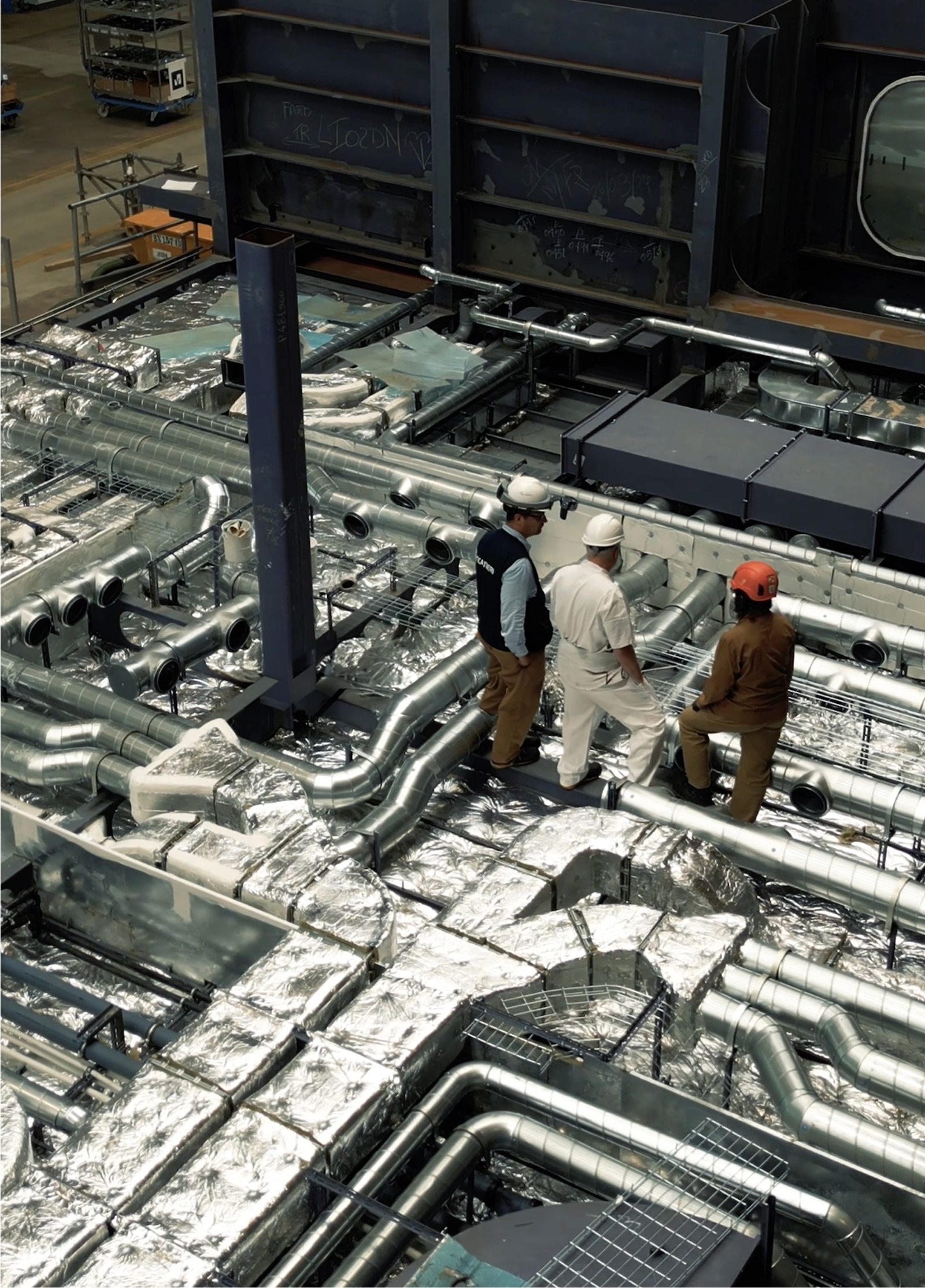 Fincantieri | Group - Sustainability - People and Community - Our People: Three people wearing helmets and work clothing on an industrial HVAC system, factory environment with metal ducts and insulated pipes, focus on technical inspection and teamwork in a professional setting.
