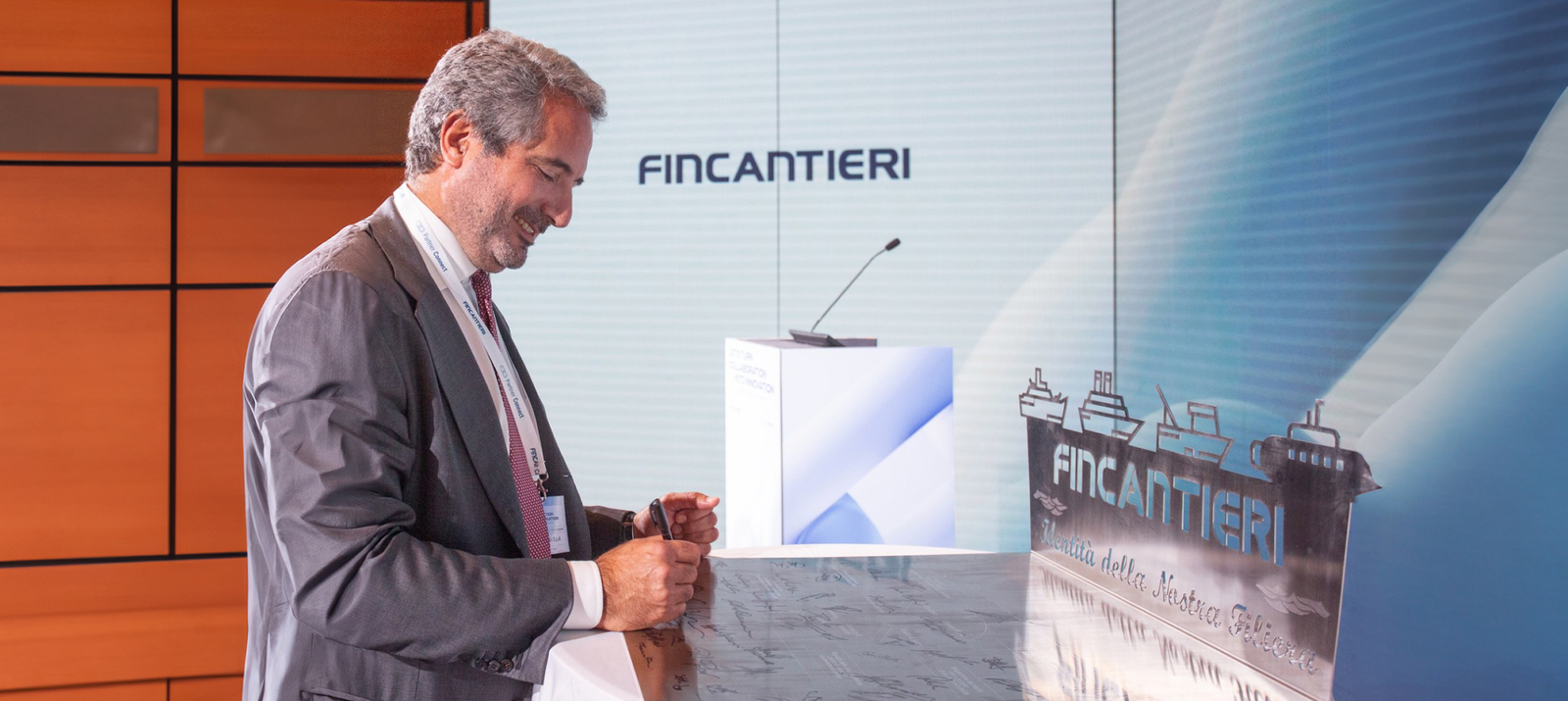 Fincantieri | Investor Relations - Suppliers and Partners - Partnership - Summit 2023: A professional setting featuring a man in formal attire near a podium with the Fincantieri logo prominently displayed. The backdrop includes maritime-themed visuals with ship illustrations and a modern design, suggesting a corporate or industry-related event.