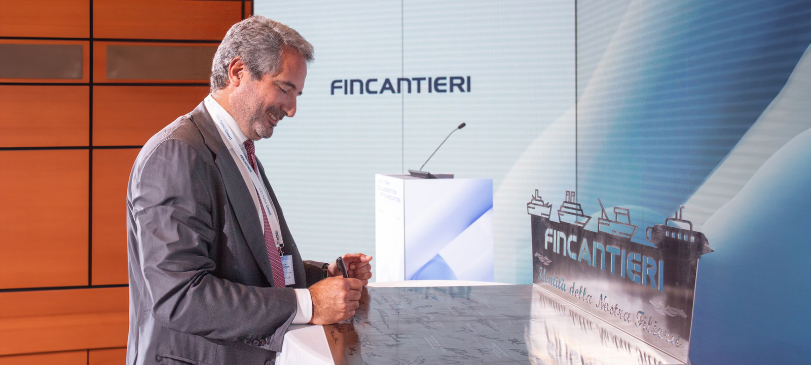 Fincantieri | Investor Relations - Suppliers and Partners - Partnership - Summit 2023: A professional setting featuring a man in formal attire near a podium with the Fincantieri logo prominently displayed. The backdrop includes maritime-themed visuals with ship illustrations and a modern design, suggesting a corporate or industry-related event.