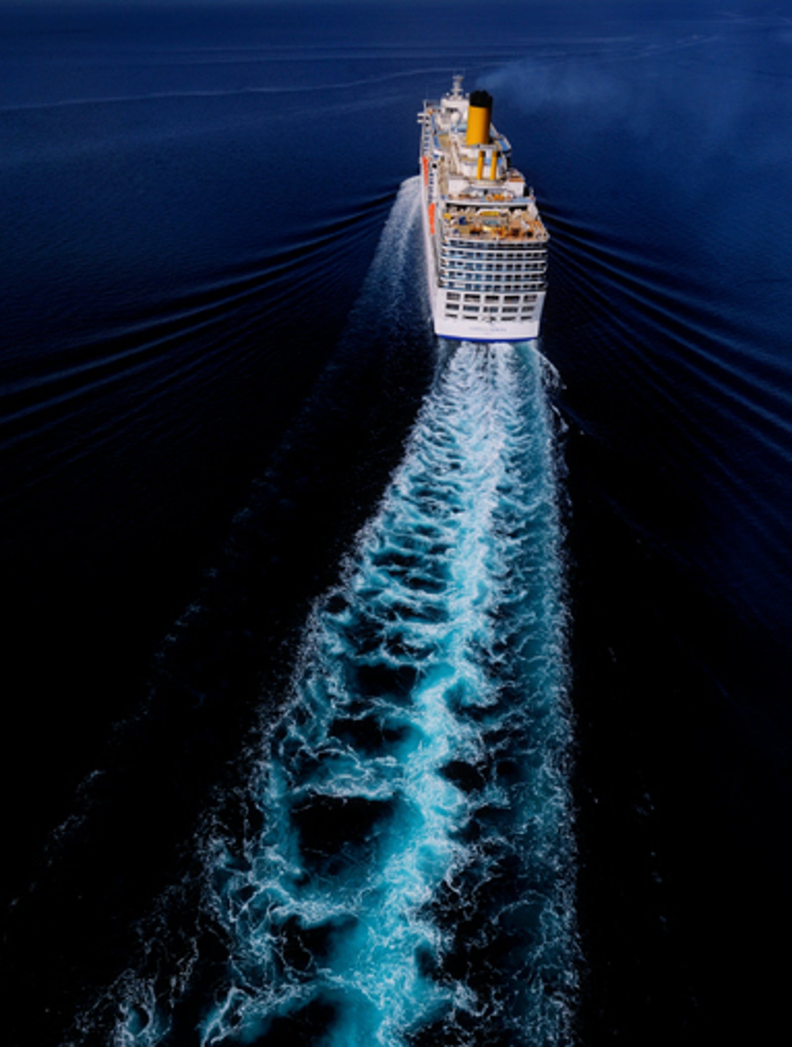 Fincantieri | Gruppo - Business - Prodotti - Navi da Crociera - Costa Luminosa: Vista aerea di grande nave da crociera operativa che naviga su oceano aperto, scia bianca evidente in contrasto con il mare blu profondo, ponti superiori e camino giallo luminoso ben visibili, scena che enfatizza movimento e scala.