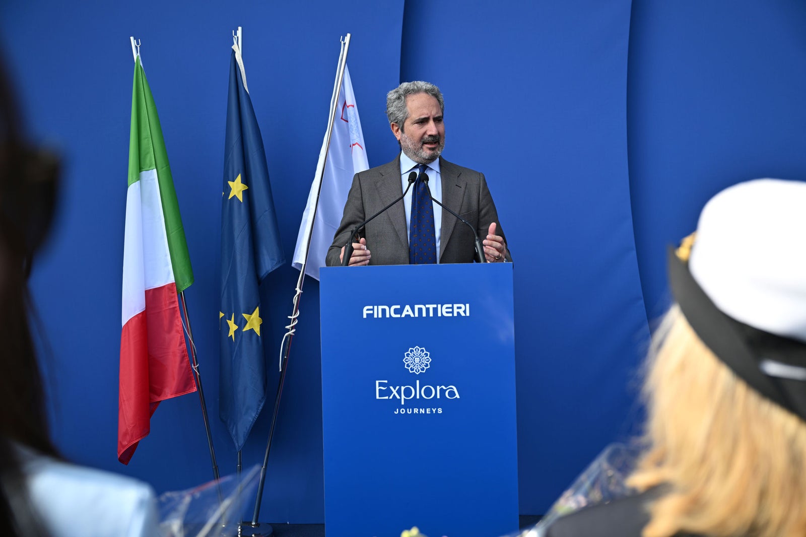 Newsroom - Press releases - 2026 - Fincantieri Group: Fincantieri:  Today triple ceremony for Fincantieri and Explora Journeys at Sestri Ponente: launche of Explora IV, coin ceremony for Explora V and steel cuttingfor Explora VI: Fincantieri executive speaks at podium with Explora Journeys logo, Italian and EU flags in background.