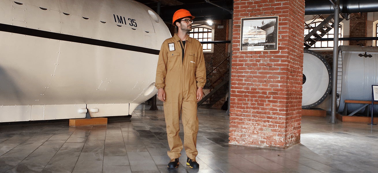 Fincantieri – People: Person wearing a beige coverall and orange safety helmet indoors near a large industrial machine labeled “IMI 35”; industrial museum setting with brick walls and exhibited machinery, lighting highlighting historical and mechanical elements.