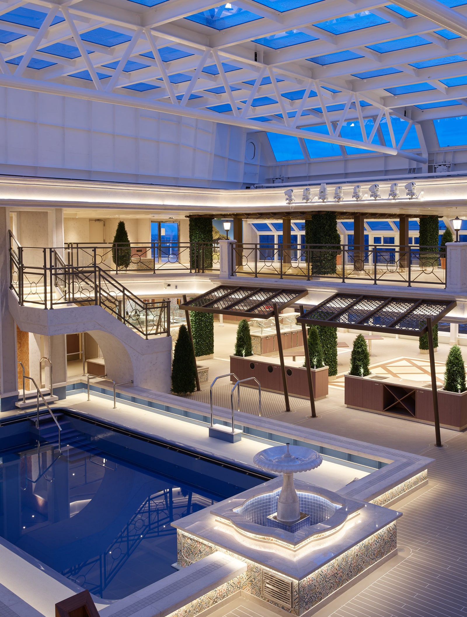 Fincantieri | Group - Business - Products - Cruise Ships - Interni Costa Firenze: Large indoor pool with elegant architecture, central fountain, decorative plants, glass ceiling filtering natural light, modern warm-tone lighting, relaxing and refined atmosphere.
