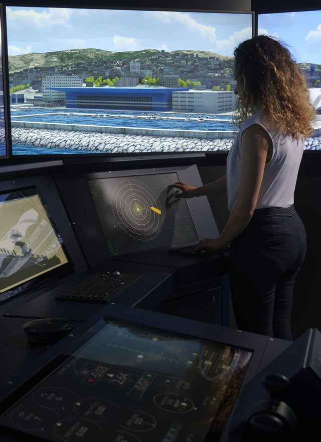 Fincantieri | Sustainability - Responsible Culture - Maritime Training: Woman at a navigation simulator in a control room, monitors displaying radar, navigation data, and coastal scenario. Detailed graphics of buildings, water, and terrain; professional environment for operational training.
