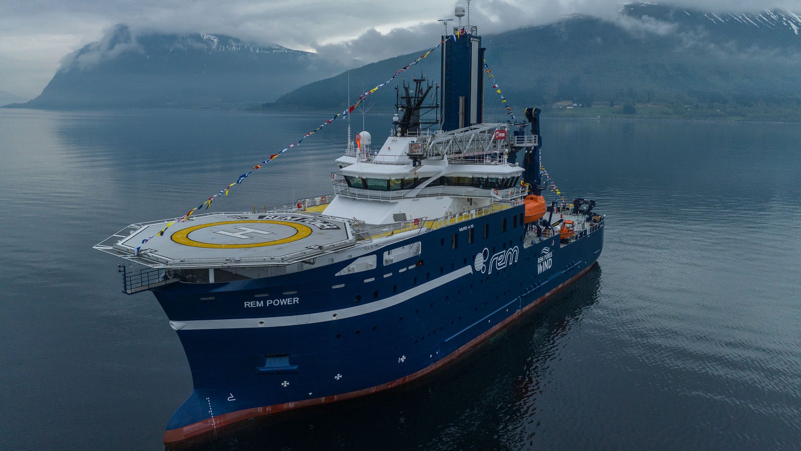 Prodotto - Offshore Support Vessel Rem Power: Vista laterale della nave operativa in un fiordo, elisuperficie con lettera H visibile, equipaggiamenti marittimi dettagliati, nome e logo Rem Power ben visibili sullo scafo, sfondo montuoso.
