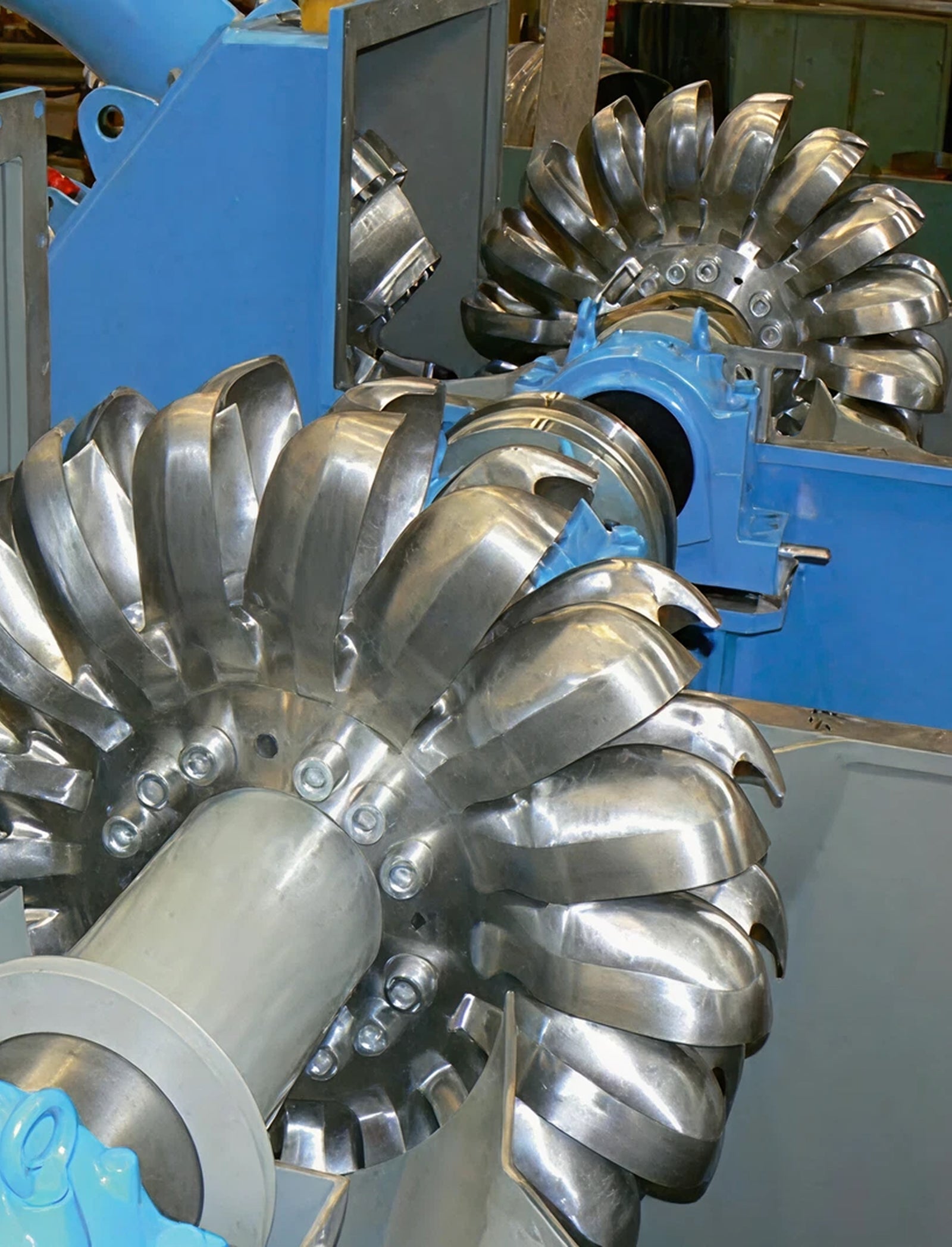 Fincantieri | Group - Subsidiaries and Affiliates - Team Turbo Machines: Detailed view of a Pelton turbine, polished metal blades engineered for high-efficiency water flow, industrial facility setting with blue structural components; highlights intricate engineering and precision of turbine design.