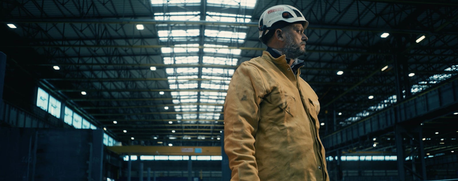 Fincantieri | Group - Sustainability - Energy Transition - Environmental Impacts: Worker wearing a helmet and protective jacket inside a large industrial warehouse, high ceilings, metal beams, and lighting from high windows. Production environment, emphasis on safety and professionalism.